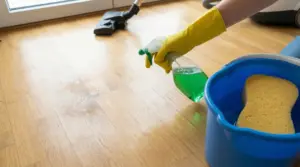 Persona con guanti gialli spruzza detergente verde su parquet accanto a spugna e secchio